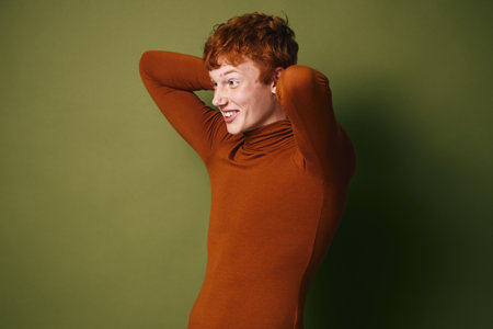 Young redhead woman with short hair wearing brown turtleneck smiling and looking to side against green background with hands behind head and relaxed poseの写真素材