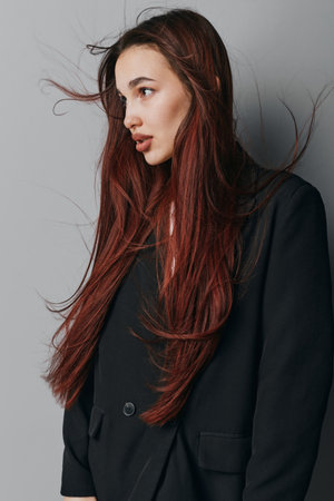 Young woman with long red hair wearing black blazer posing sideways against gray background. Natural look with loose hair strands and casual style in studio portrait photography.の写真素材