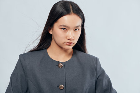 Portrait of confident young woman with long black hair wearing gray blazer posing serious and stylish on light background. Female model with natural look and professional attitude indoors.の写真素材