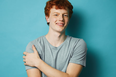 Young man with red hair and casual gray t shirt standing smiling on blue background. Male portrait with confident expression and relaxed posture in studio environment.の写真素材