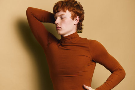 Young man with red hair in brown turtleneck posing with eyes closed and hand behind head against...