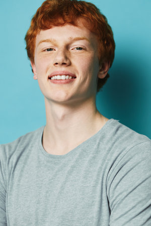 Teenager with red hair and smiling face wearing gray t shirt against blue background. Portrait of...
