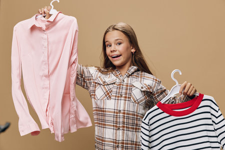 Girl with long hair holding two clothes hangers with shirt and sweater, smiling and wearing plaid shirt on brown background. Fashion choice, style and clothing options for kids.の写真素材
