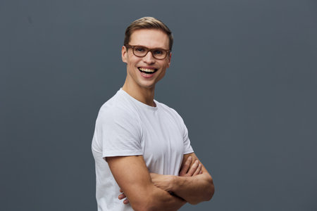 Smiling happy young Caucasian man wearing glasses and a white t-shirt, looking at camera with friendly expression, arms crossed, casual pose, studio background, lifestyle concept.の写真素材