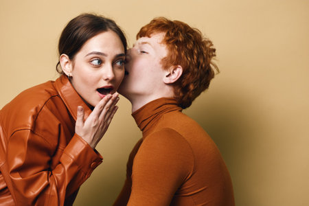 Woman shares a secret whisper to surprised friend with hand covering mouth on beige background. Two...