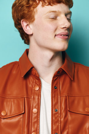 Young man with red hair and freckled skin wearing brown leather jacket and white t shirt eyes closed on blue background. Portrait of peaceful male with stylish casual look and calm expression.の写真素材