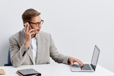 Businessman in gray blazer and glasses working at laptop while talking on mobile phone isolated on plain white background. Office workspace concept, professional, concentration, technology.の写真素材