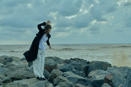 Woman on rocky shore battling wind by the sea, wearing a white dress and dark coat, dramatic clouds overhead, movement captured as waves surge and the horizon opens into distanceの写真素材