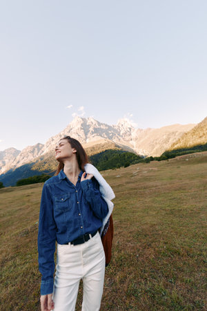 Woman stands in a meadow with mountains behind, outdoors portrait highlighting fashion outfit and...