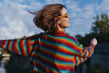 Woman in a rainbow sweater and sunglasses, smiling with arms outstretched outdoors, short hair and casual outfit enjoying a windy urban rooftop scene, joyful travel lifestyle moment.の写真素材