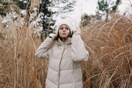 Woman in a beige puffer jacket stands among tall dry grasses in a chilly outdoor scene, conveying...