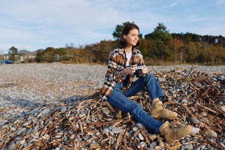 Woman outdoors with camera sits on a leaf pile near a pebble beach, autumn light, casual plaid shirt, blue jeans and boots, calm creative mood for nature photographyの写真素材