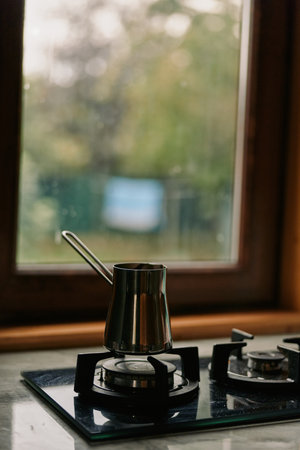 Coffee pot on stovetop in a cozy kitchen by a window, stainless metal pot on a gas stove burner,...