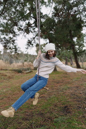 Girl on a wooden swing in an outdoor setting, wearing a knit sweater, beanie and jeans, enjoying a...