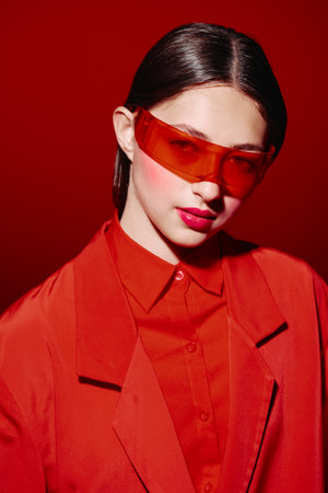 Young woman in vibrant red outfit and transparent orange sunglasses poses against red background....