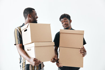 Two african american men carrying cardboard boxes for moving or delivery, teamwork and cooperation with casual clothes on white background, young black men holding packages together.の写真素材
