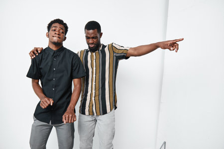 Two young black men with dark skin posing together in casual clothes, one pointing and the other smiling with eyes closed on white background. Friendship and style concept.の写真素材