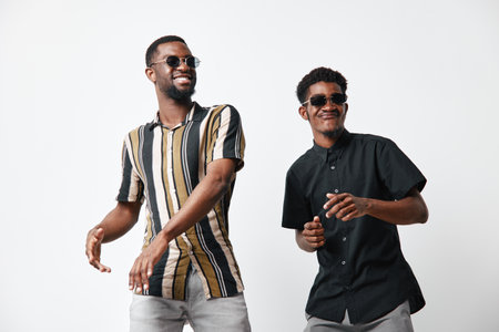Two young black men with dark skin wearing sunglasses and casual shirts smiling and dancing together on white background for friendship and lifestyle.の写真素材