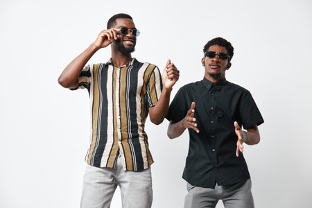 Two African American men with dark skin wearing sunglasses and casual shirts dancing and enjoying music together on white background in studio. Friends having fun and smiling.の写真素材