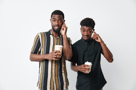 Two young African men with dark skin holding coffee cups and pointing to their heads on white...