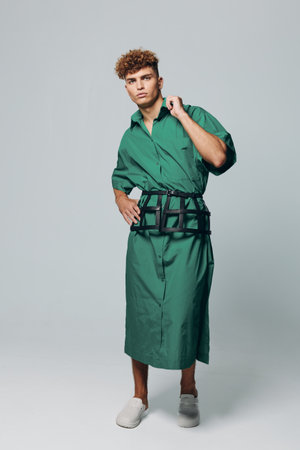 Young man with curly hair wearing green long shirt dress and black belt bag posing in studio with neutral background. Casual fashion, confident model and modern street style outfit.の写真素材