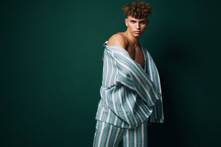 Young man with curly hair wearing striped pajamas posing against dark green background, fashion...