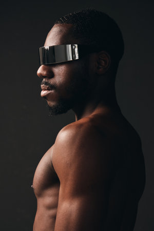 Creative visual designer style portrait of black man with dark skin wearing futuristic reflective visor glasses in studio. Side view, bare torso and modern fashion concept with minimal lighting.の写真素材