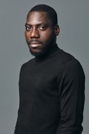 African man portrait in creative conceptual visual style wearing black turtleneck sweater against gray background. Confident young male model with short hair and beard looking at camera.の写真素材