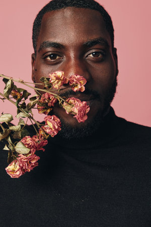 African man with dark skin holding dried pink flowers close to face in creative conceptual visual style. Portrait with black turtleneck and soft pink background for artistic expression.の写真素材