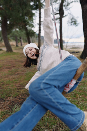 Girl on a swing in an outdoor park, laughing with joy as she soars, cozy sweater and jeans, trees in...