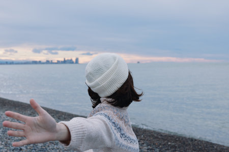 Woman standing on a pebble beach with outstretched arms, wearing a white beanie and knitted sweater,...
