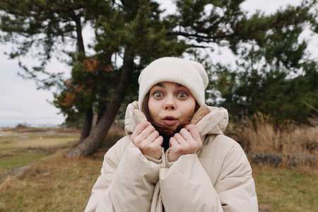 Woman in a beige winter coat and beanie outdoors, surprised expression as she pulls the collar tight...