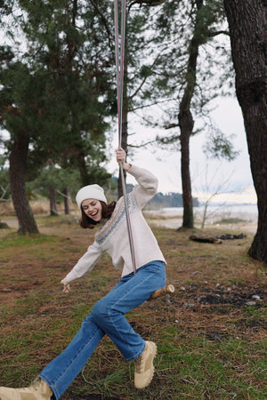 Outdoor swing moment with a girl in a knit sweater and jeans swinging beside trees by a lakeside,...