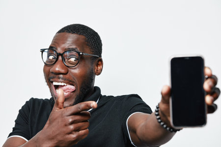 African American man with dark skin and glasses smiling and pointing at smartphone with blank screen in hand on white background. Casual clothing and happy expression with tech device.の写真素材