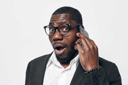 African American man with dark skin wearing glasses and black suit talking on smartphone with surprised expression on white background. Communication, technology and business concept.の写真素材