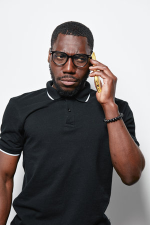 Black man with dark skin wearing glasses and black polo shirt talking on smartphone with serious...