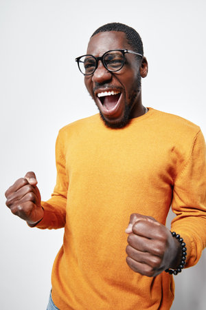 African American man with dark skin wearing glasses and orange sweater expressing excitement with clenched fists and open mouth on white background. Happy, energetic, casual style.の写真素材