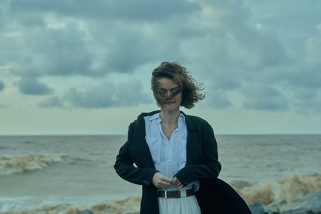 Business woman on a windy shoreline adjusts her coat as waves crash, projecting resilience, coastal...