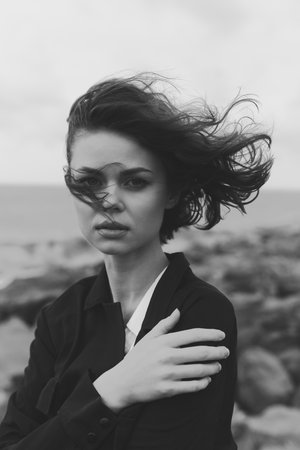Woman in a blazer stands against a windy seaside backdrop, monochrome tones and moody atmosphere highlighting her expression and outdoor styleの写真素材