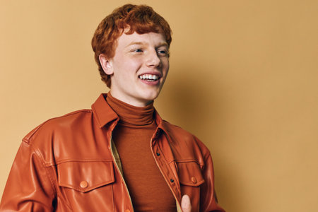 Young man with red hair smiling in brown leather jacket and turtleneck standing against beige...