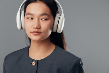 Young woman wearing white headphones and dark jacket looks aside on gray background. Portrait of confident girl with long hair listening to music or audio in casual style.の写真素材
