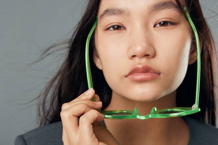 Young woman with long dark hair holds green glasses close to her face in closeup portrait. Serious expression with natural makeup and gray background in studio light.の写真素材
