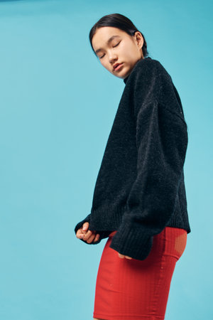 Young woman wearing black sweater and red skirt standing sideways with eyes closed on blue background. Fashion model in casual outfit with relaxed expression and minimal makeup indoor.の写真素材