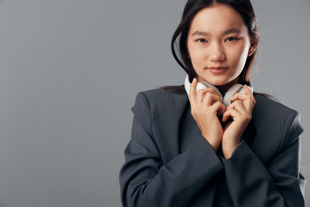 Young woman with headphones holding them close to face against gray background. Portrait of confident female in dark jacket looking at camera, modern style with natural expression and minimal makeup.の写真素材