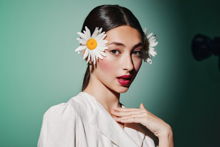 Young woman with smooth skin wearing white blouse poses with daisies in hair against a green background. Creative fashion beauty portrait with a fresh and natural vibe.の写真素材