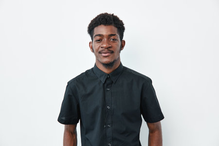 Young African man with dark skin wearing black short sleeve shirt standing and smiling on white background. Portrait of confident male with natural hair and casual style.の写真素材