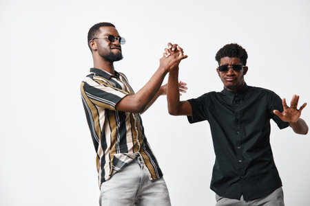 Two young black men with dark skin wearing sunglasses and casual shirts dancing and having fun together on white background in studio.の写真素材