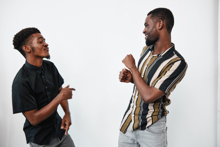 Two men with dark skin dancing and smiling together in casual clothes against white background. Friends enjoying music and having fun in relaxed atmosphere indoors.の写真素材