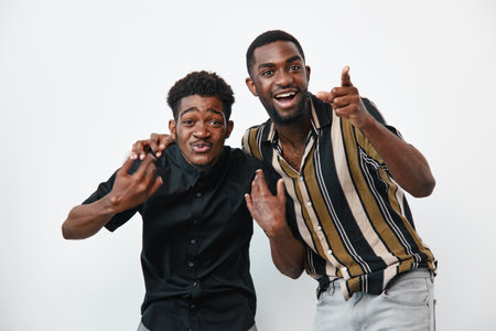Two young black men with dark skin posing together on white background, smiling and making hand gestures, casual clothing and joyful expressions in studio.の写真素材