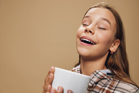 Young woman with closed eyes enjoying aroma of hot coffee or tea in hands wearing casual plaid shirt on beige background with peaceful expression and smile.の写真素材